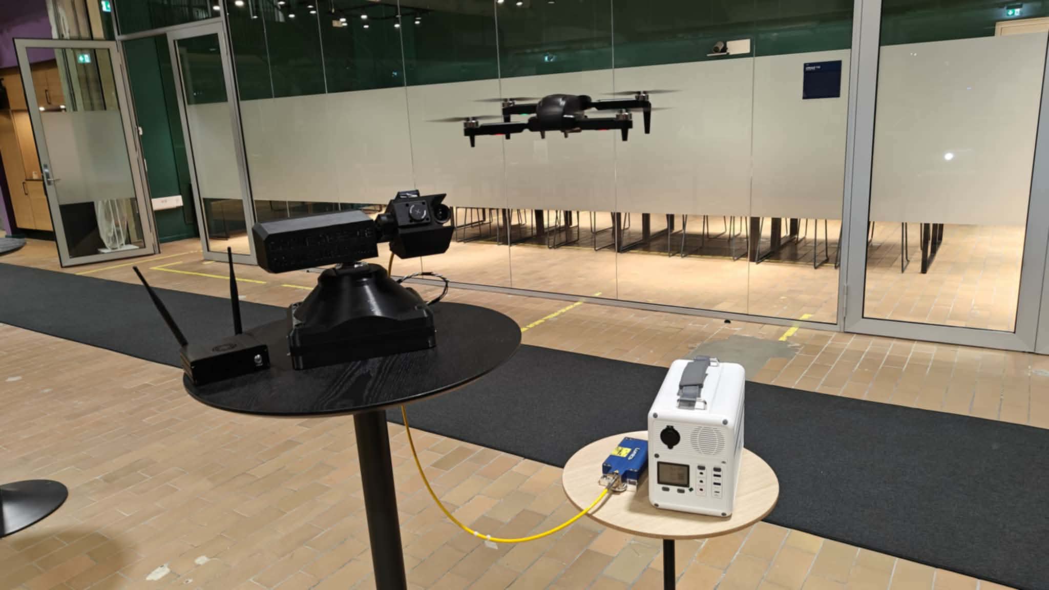 The turret indoors on a stand, holding track on a small quadcopter hovering across the hall, with a portable battery unit connected by a yellow fibre.